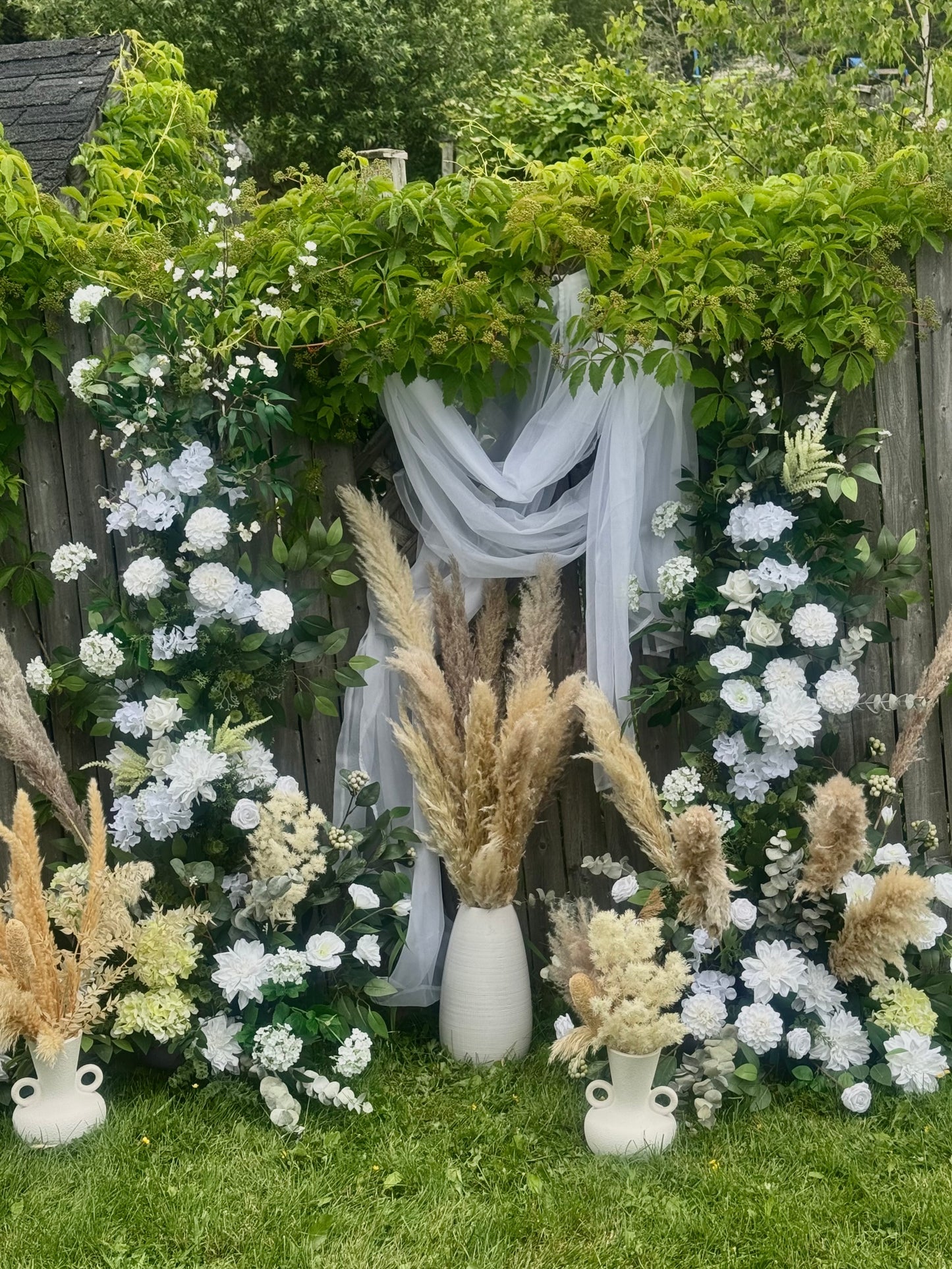 Garden style outdoor setup with flowers, pampas grass, and white fabric against a wooden fence.